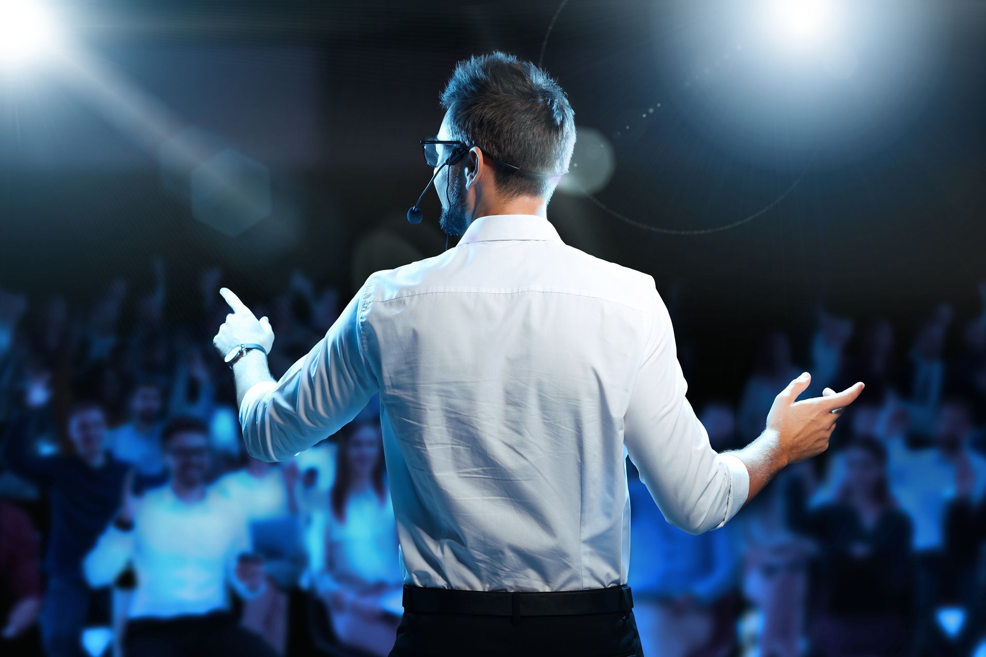Man speaking at a large event, gesturing with hands. Audience in background, illuminated by blue light.
