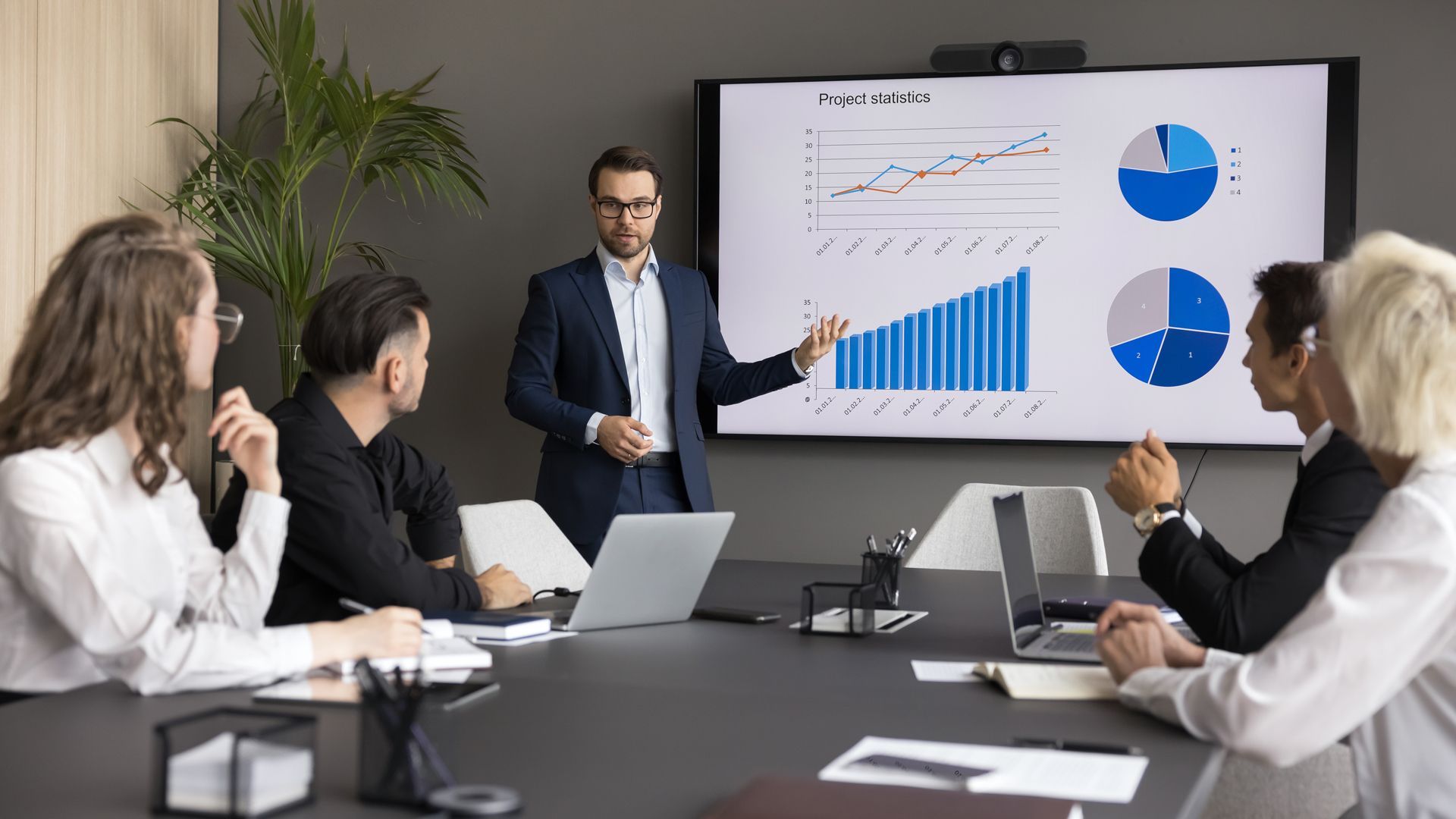 Man in suit presents charts on a screen to a team in a meeting room.