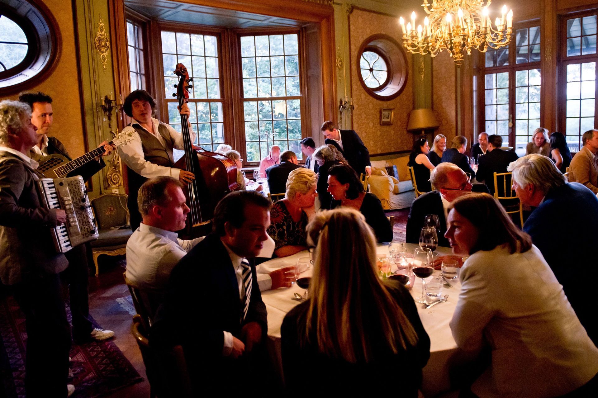 In de sfeervol verlichte, sierlijke eetzaal met grote ramen en een kroonluchter speelt een band voor de gasten die aan het eten zijn.