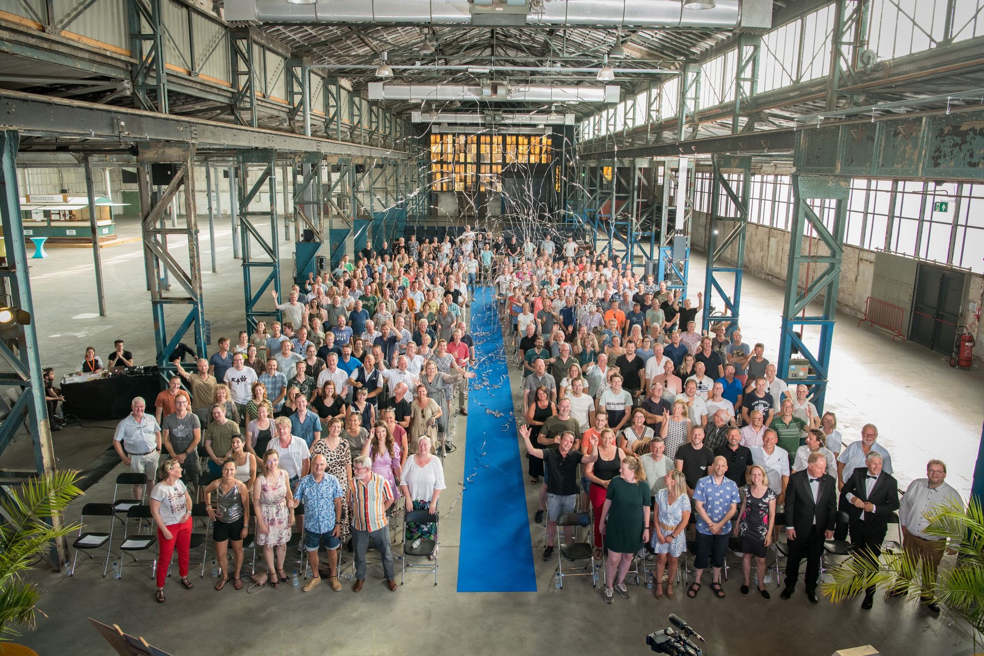 Een grote menigte was binnen verzameld voor een evenement; blauwe loper, industriële architectuur, veel mensen keken naar de camera.
