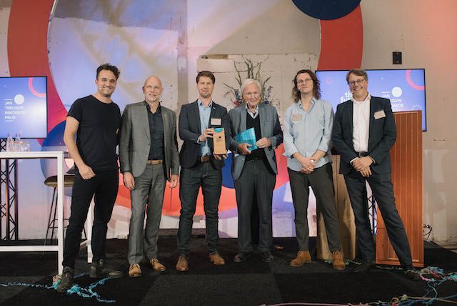 Een groep van zeven personen poseert met prijzen op een podium met schermen en een kleurrijke achtergrond.