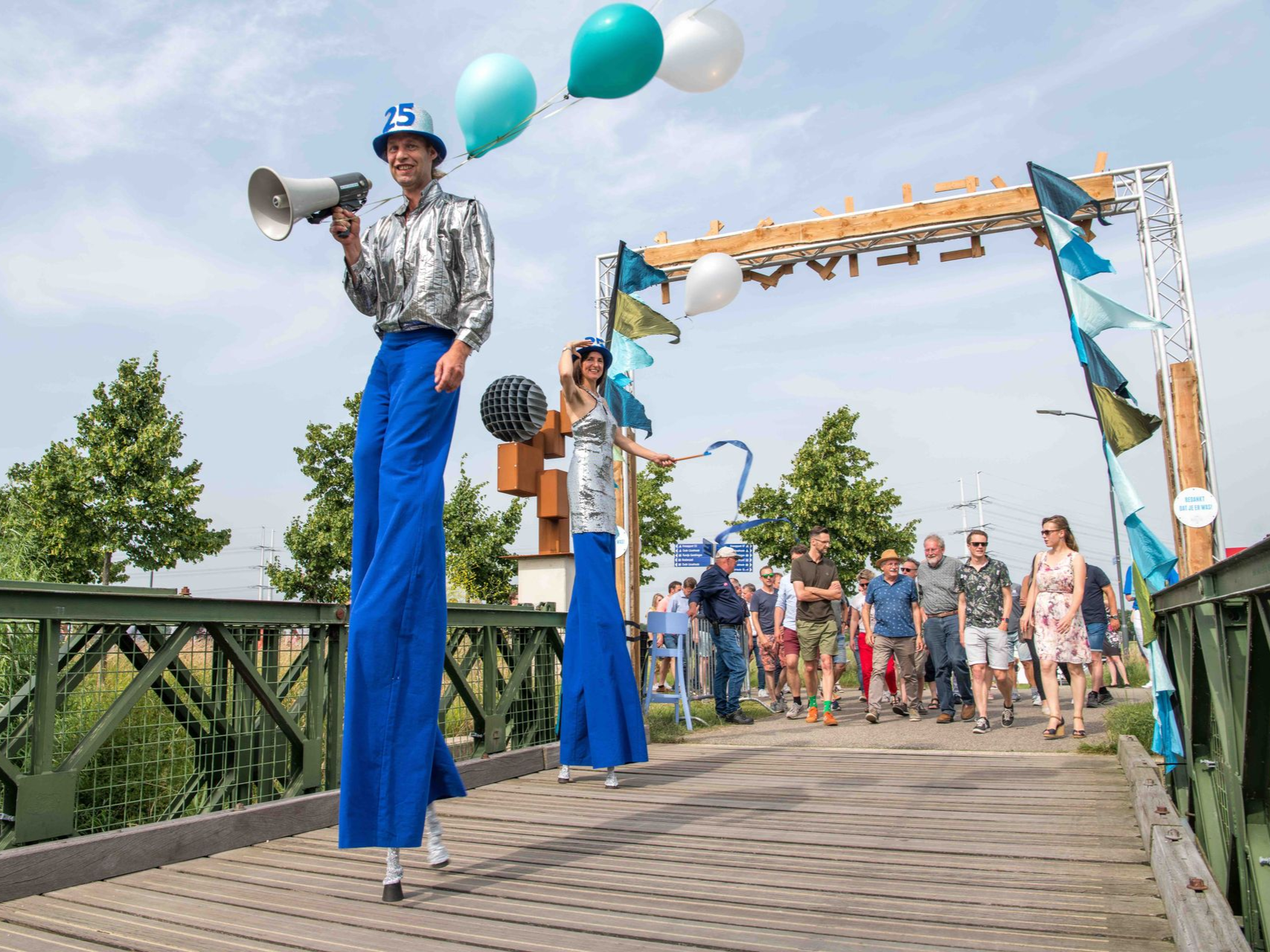 Twee mensen op stelten met een megafoon en linten leiden een parade op een brug onder een boog, met omstanders.