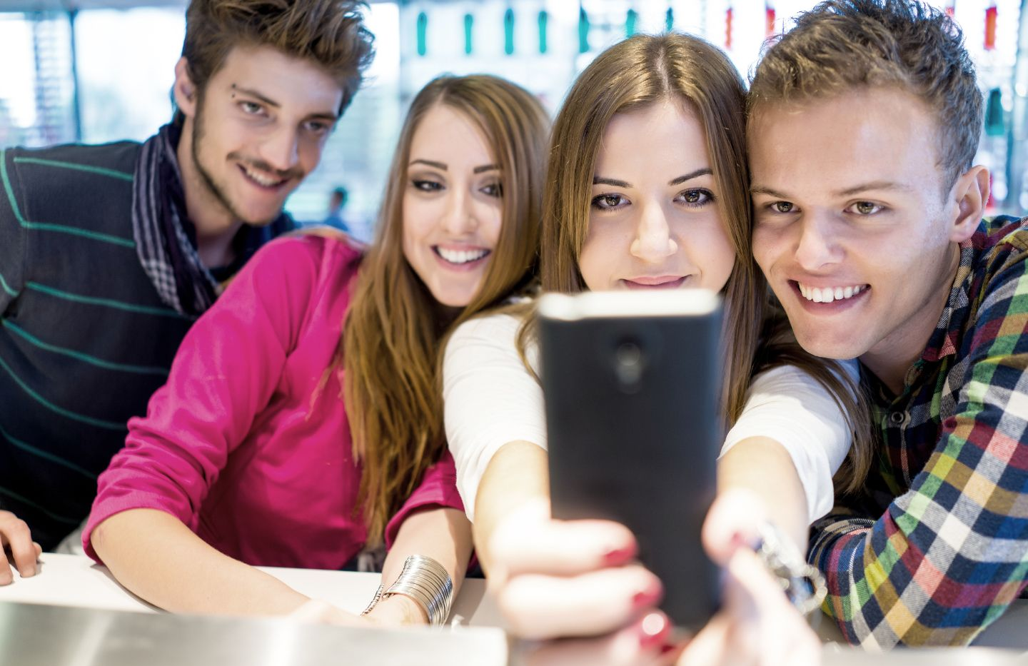 Vier jongeren lachen naar een telefoon en nemen een selfie. Binnen, fel licht, verschillende casual outfits.