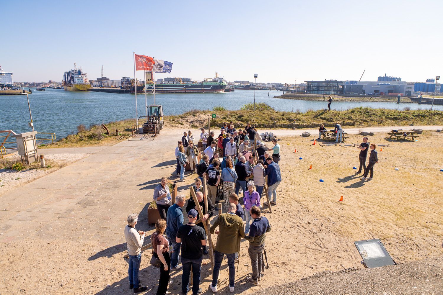 Een grote groep mensen verzamelde zich op een zonnige dag bij een waterpartij. Sommigen hielden stokken vast, met vlaggen op de achtergrond.