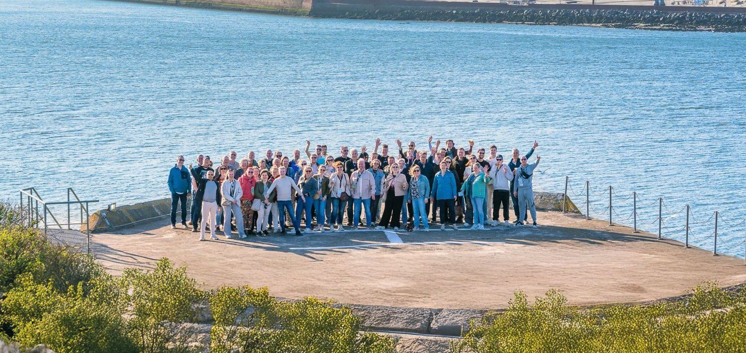 Een groep mensen staat op een zanderig platform bij het water, velen met opgeheven armen. Het is een stralende, zonnige dag.