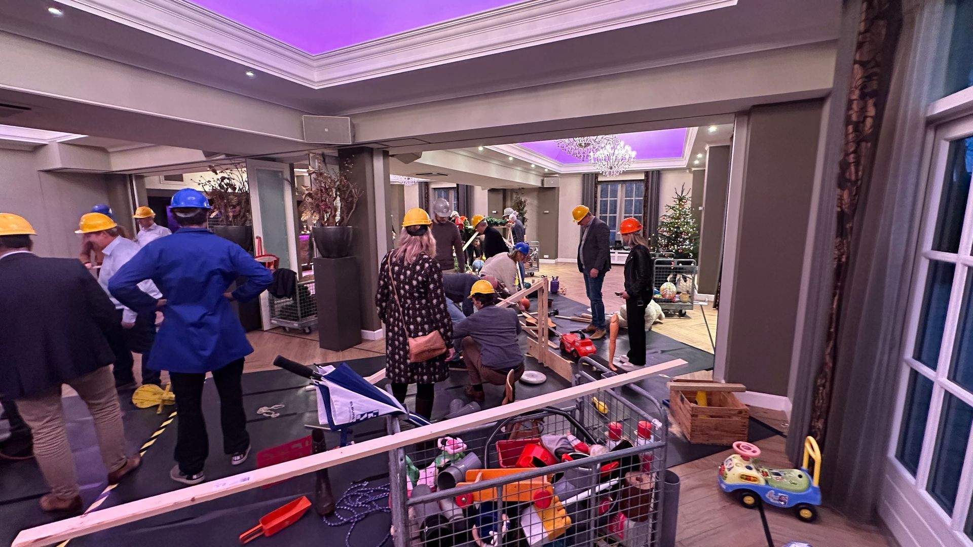 Mensen met helmen in een gebouw met bouwmachines. Sommigen kijken naar het plafond.