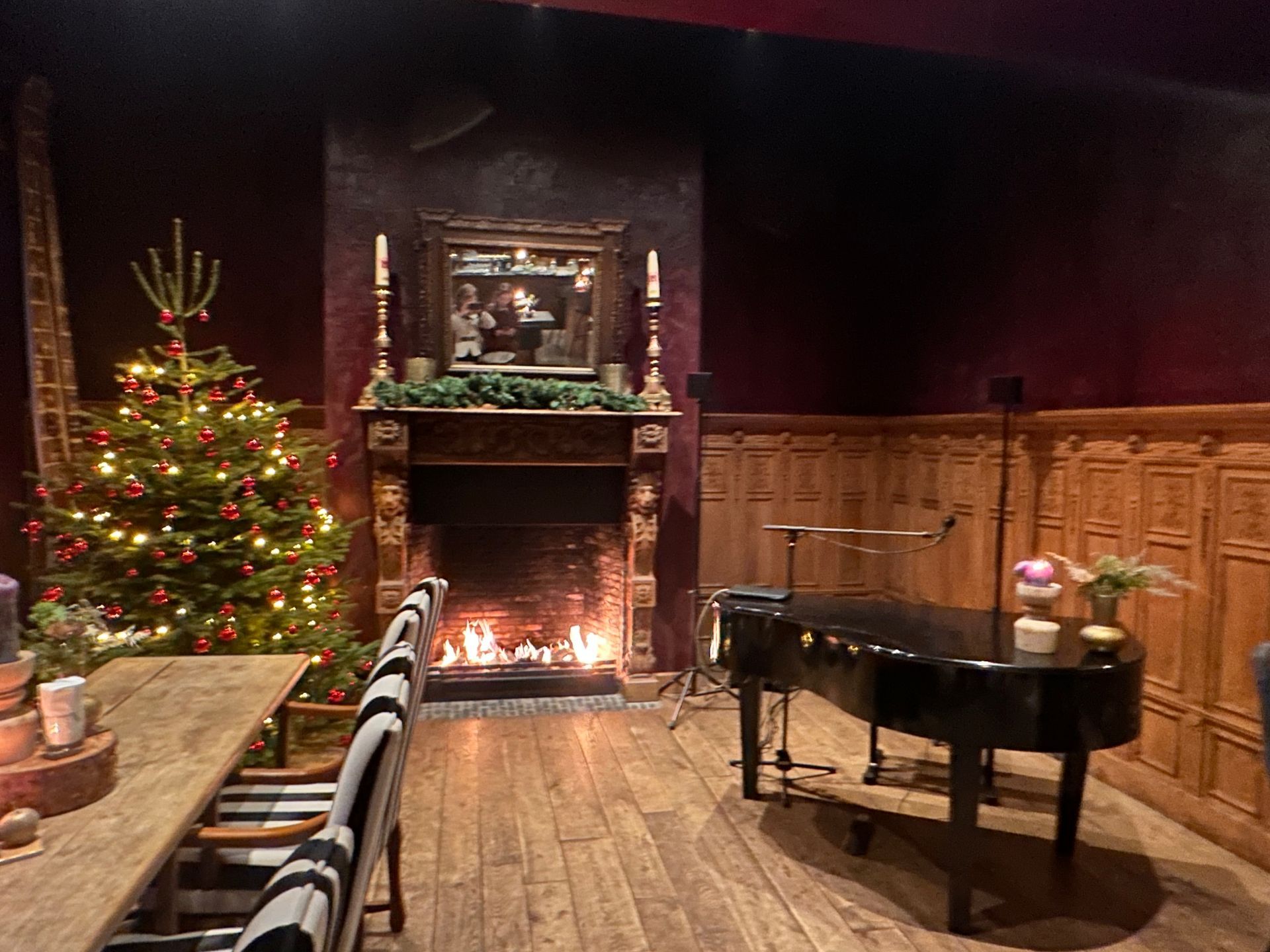 Gezellige kamer met kerstboom, open haard, piano en houten lambrisering.