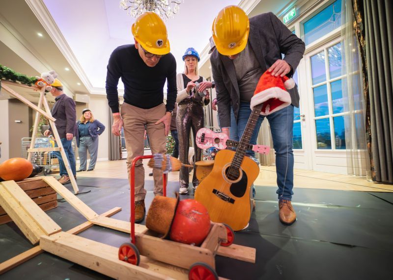 Mensen met bouwhelmen bekijken een houten apparaat met een gitaar en een kerstmuts in een versierde kamer.