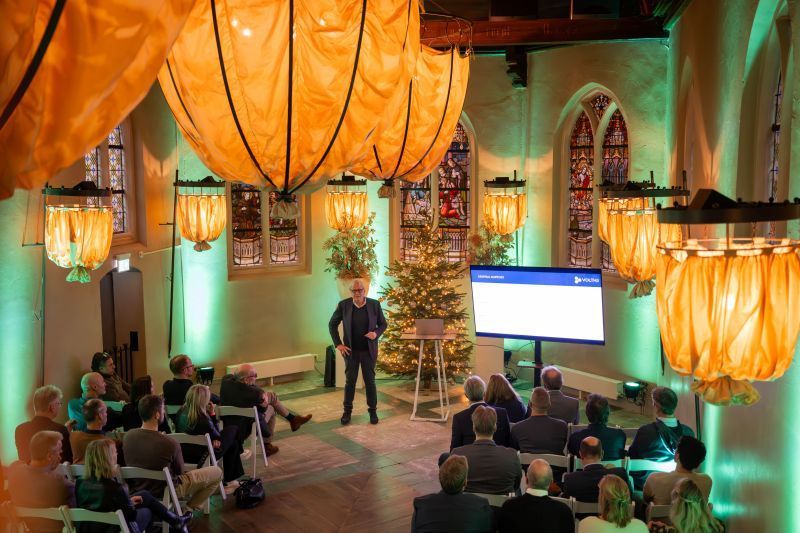 Een spreker geeft een lezing voor het publiek in een versierde kerkachtige ruimte, met hangende lantaarns en een kerstboom.