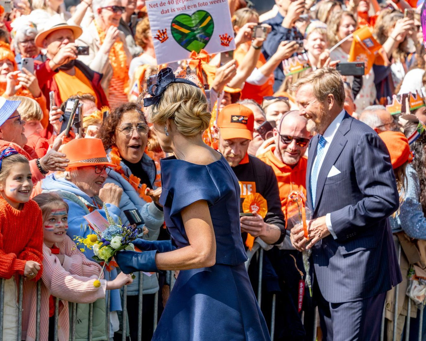 De Nederlandse koninklijke familie begroet een menigte; de ​​koningin in het blauw neemt bloemen in ontvangst, de koning in pak glimlacht. Mensen dragen oranje, zwaaien en juichen.