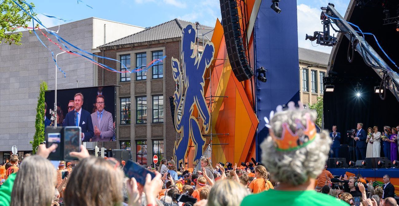 Het publiek bekijkt het evenement op het podium met een oranje achtergrond en een scherm waarop mensen te zien zijn. Confetti, daglicht.