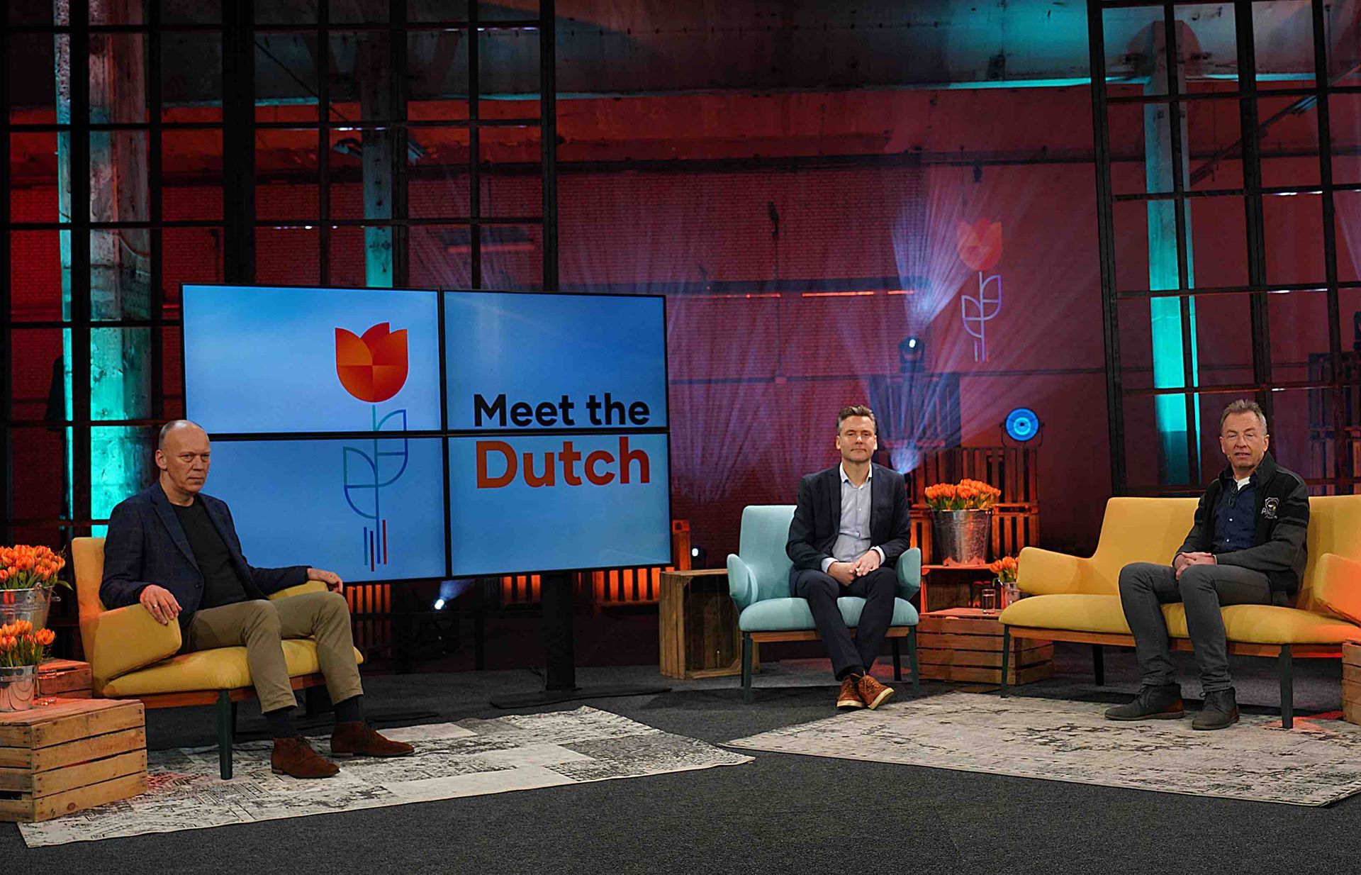 Drie mensen zitten op een podium, voor een scherm met het logo van