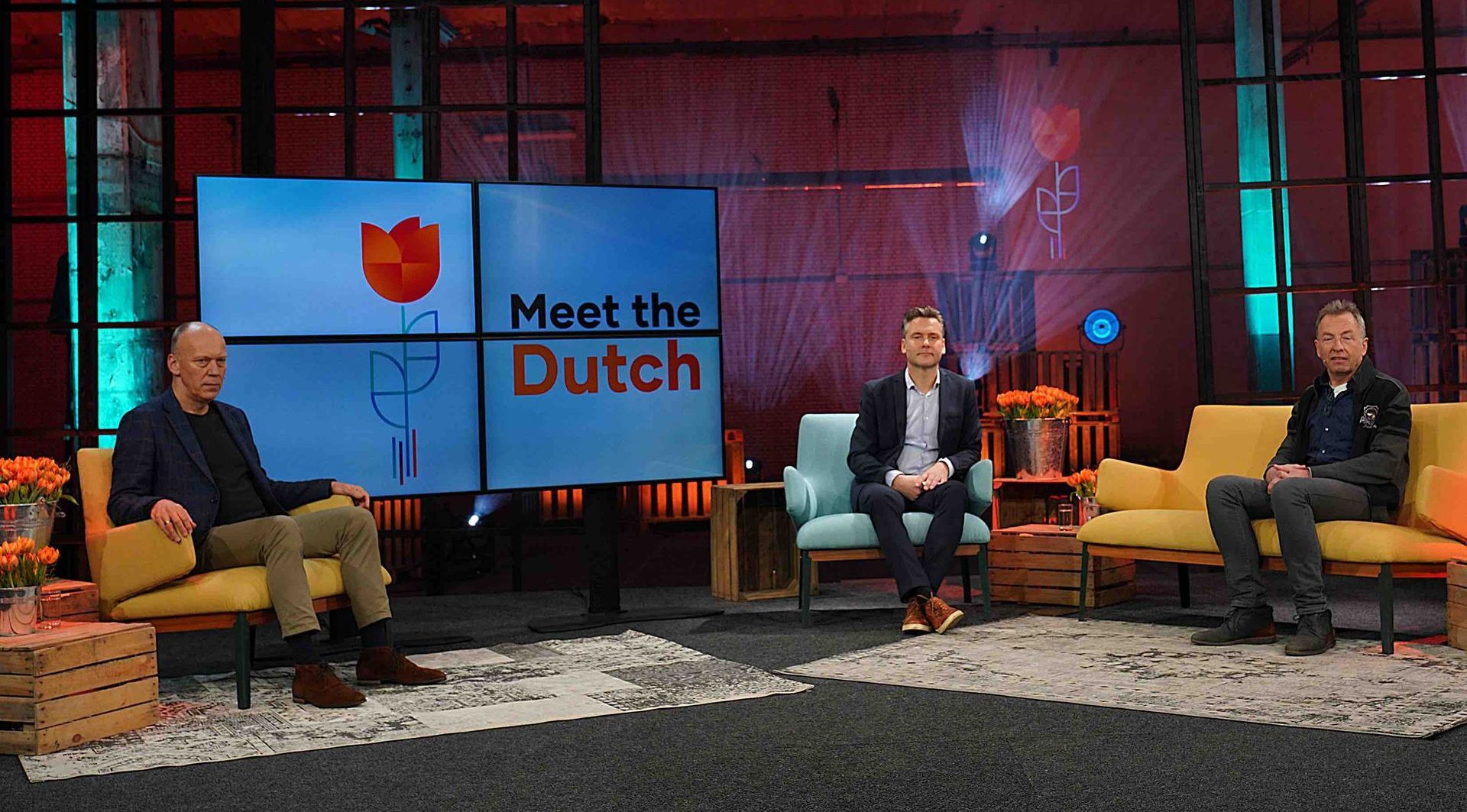 Drie mannen op een podium voor een show met de titel "Meet the Dutch". Twee mannen zitten op banken, één in het midden met een microfoon.