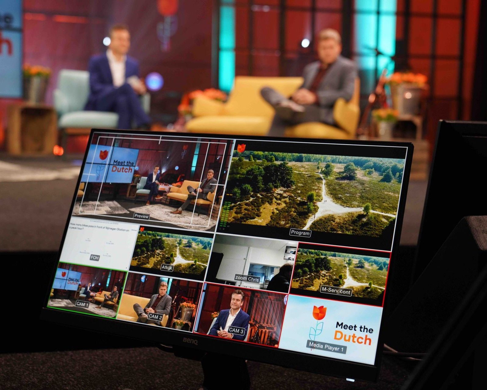 Een live tv-showset met twee presentatoren. Een monitor toont verschillende camerahoeken en een logo.