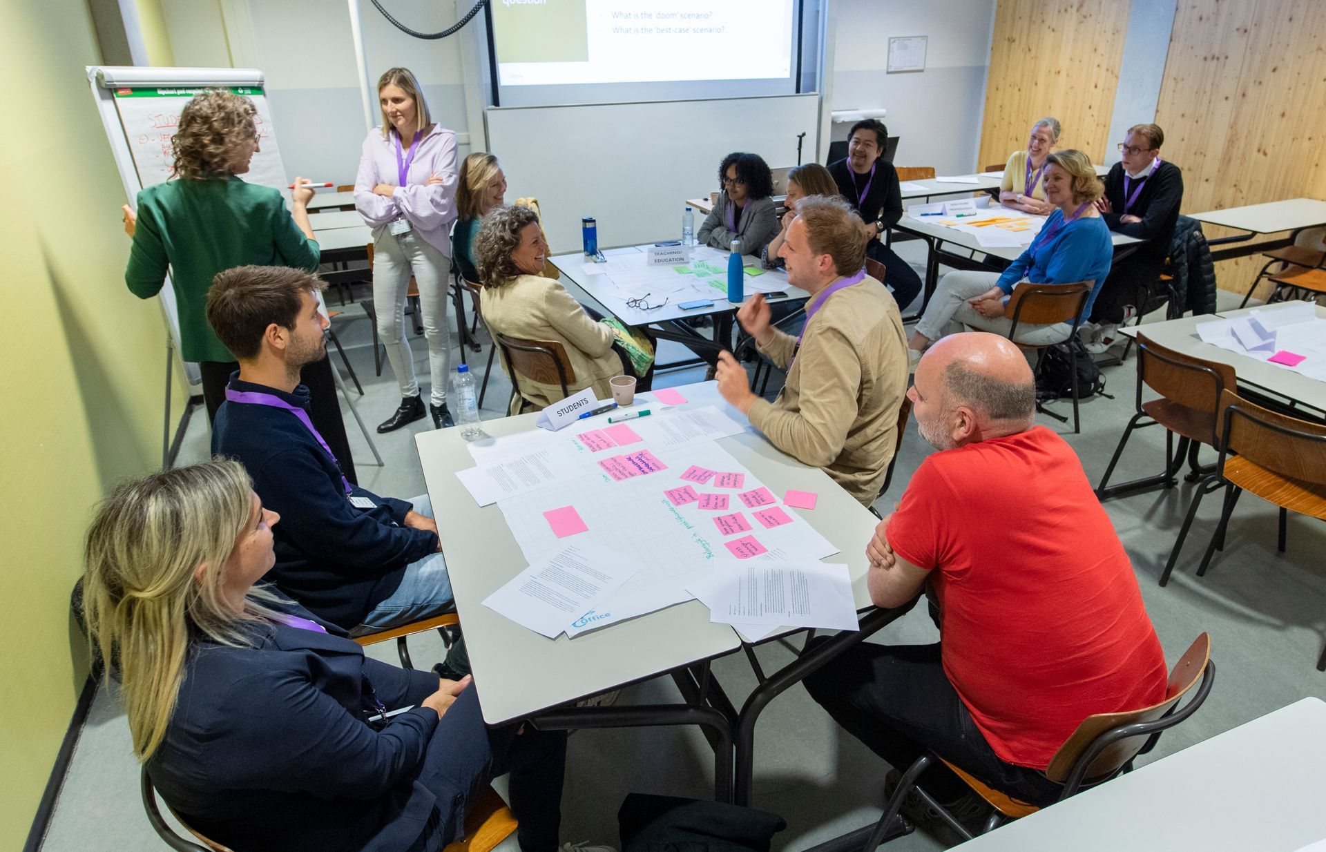 Een groep mensen in een vergadering, rond tafels met papieren. Sommigen praten, anderen kijken toe. Een presentator staat bij een whiteboard.