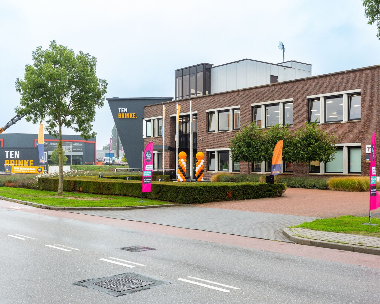 Buitenaanzicht van een bakstenen gebouw met ramen, groene heggen en een grijze weg. Het bordje 'TEN HUURBAAR' is zichtbaar.