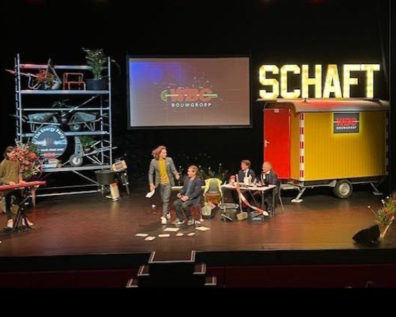 Podiumdecor met artiesten en een trailer. Op een scherm is 