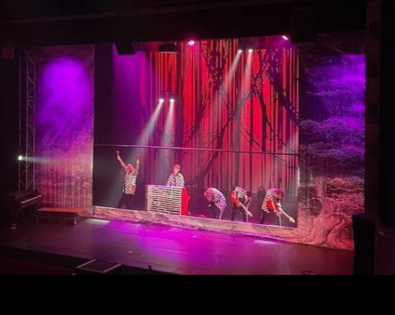 Podium met acteurs onder paars en rood licht. Artiesten in shirts en broeken met print.