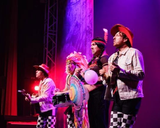 Band op het podium in kleurrijke kostuums, die instrumenten bespelen en onder toneelverlichting staan.
