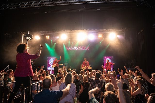 Een band treedt op op het podium, het publiek juicht met de armen omhoog en er zijn felle lichten op het podium.