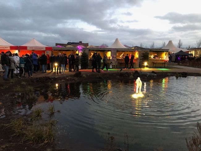 Mensen op een kerstmarkt bij een vijver met verlichte versieringen en kraampjes onder een bewolkte hemel.