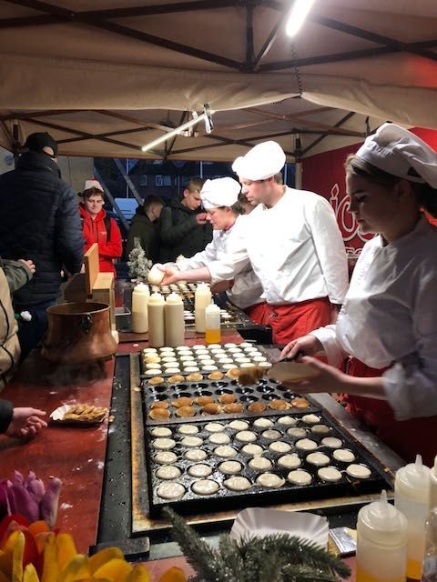 Chefs bakken kleine pannenkoekjes bij een marktkraam. Ze dragen witte hoeden en schorten, te midden van een menigte mensen.