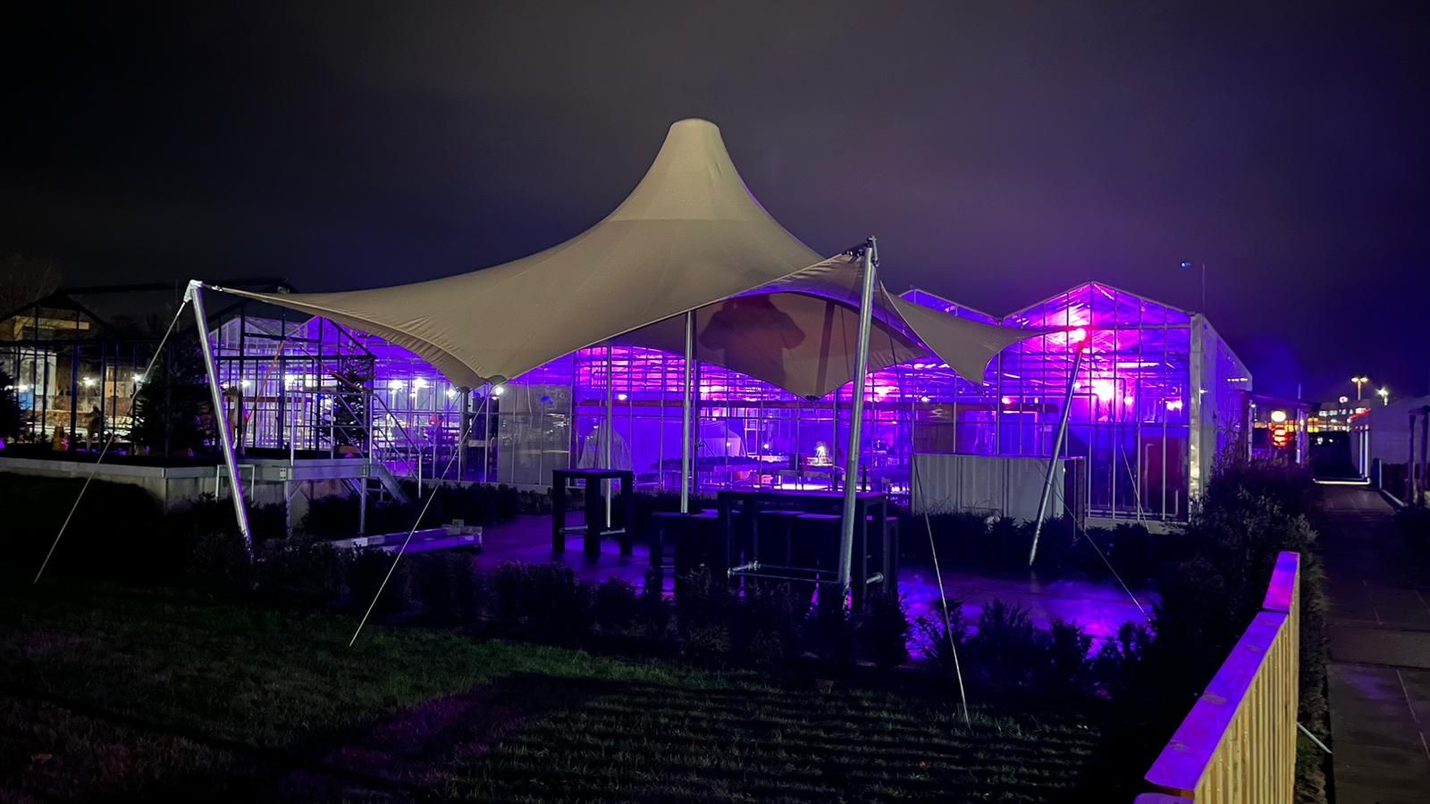 Een restaurant met een tentvormig dak dat 's avonds verlicht is met paarse lampjes.