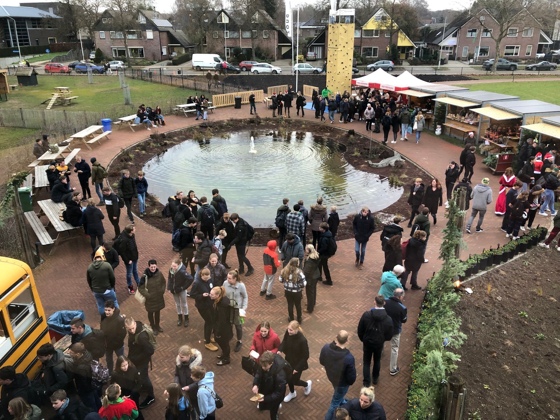 Mensen verzamelden zich rond een vijver met een fontein, kraampjes en een klimwand tijdens een buitenevenement.