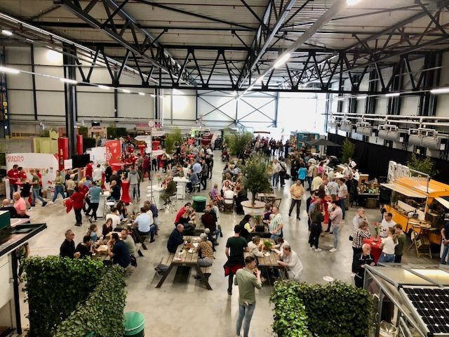Groot indoorevenement met veel mensen, eetkraampjes, tafels en decoraties onder een hoog plafond.