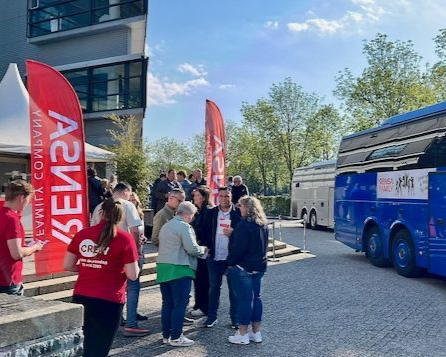 Mensen verzamelen zich buiten een gebouw, rode vlaggen, blauwe bus. Iemand draagt een rood