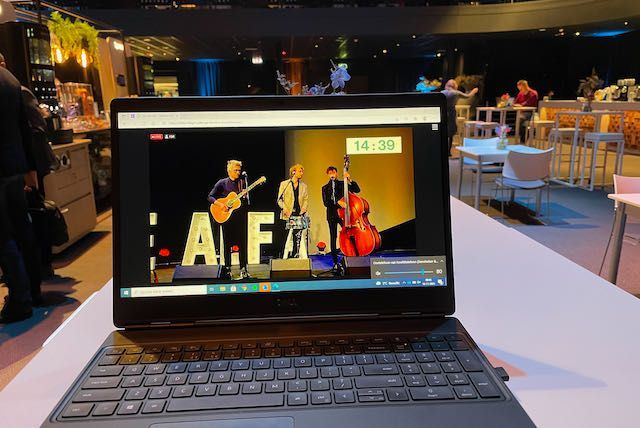 Laptop met een livestream van een band die op een podium optreedt. De laptop staat op een wit oppervlak in een bar.