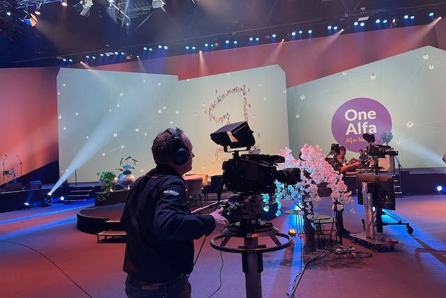 Een cameraman filmt op een podium met een achtergrond, een logo en artiesten.