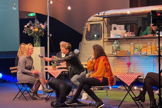 Drie mensen zitten aan een tafel, waarvan één een microfoon vasthoudt. Op de achtergrond staat een foodtruck.