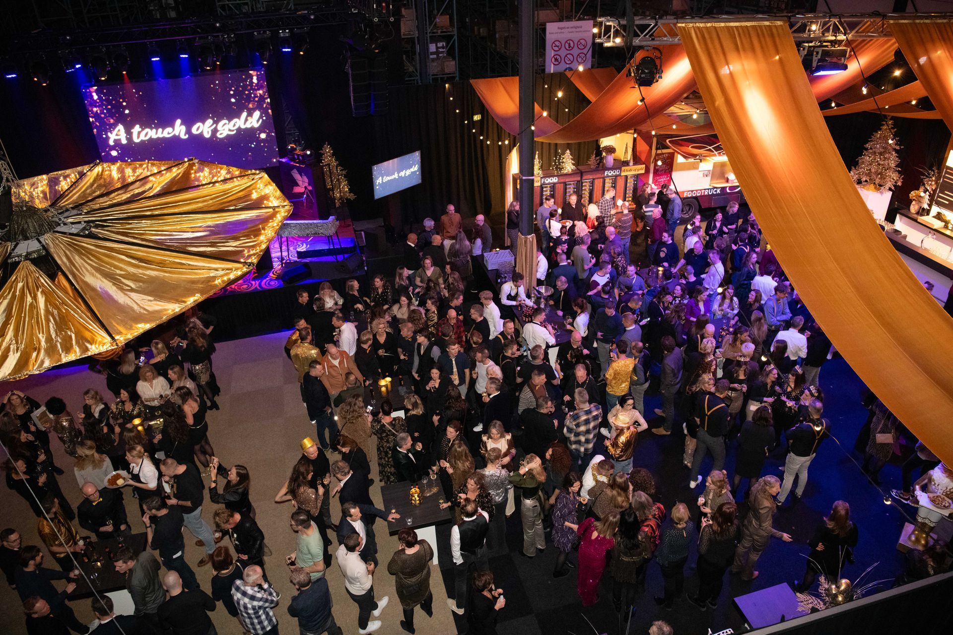 Groot evenement met publiek, podium en gouden decoratie. Mensen mengen zich en staan in een grote open ruimte.