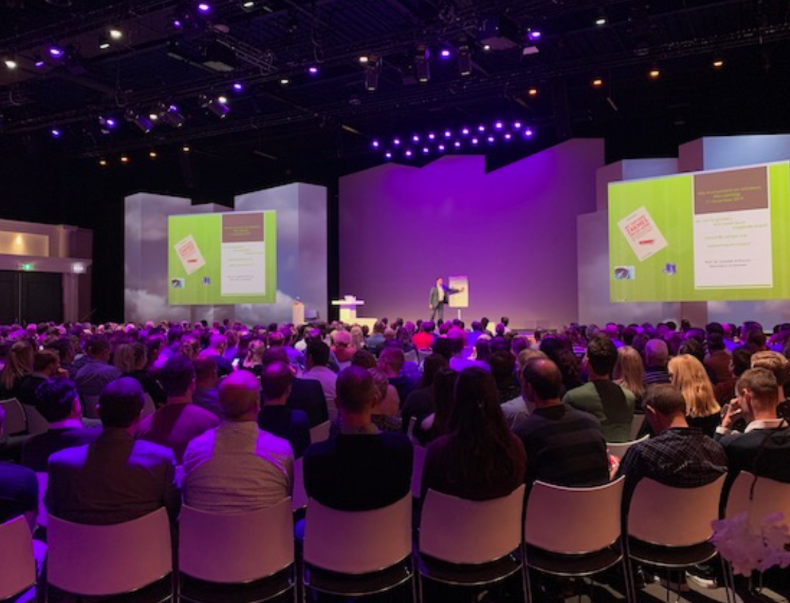 Publiek op stoelen, kijkend naar een podium met een spreker en presentatieschermen. Paarse verlichting.