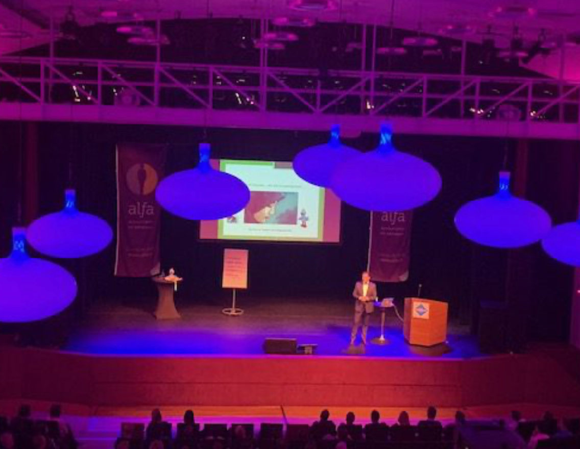 Een man spreekt op een conferentiepodium, met blauwe plafondlampen. Het publiek zit vooraan.