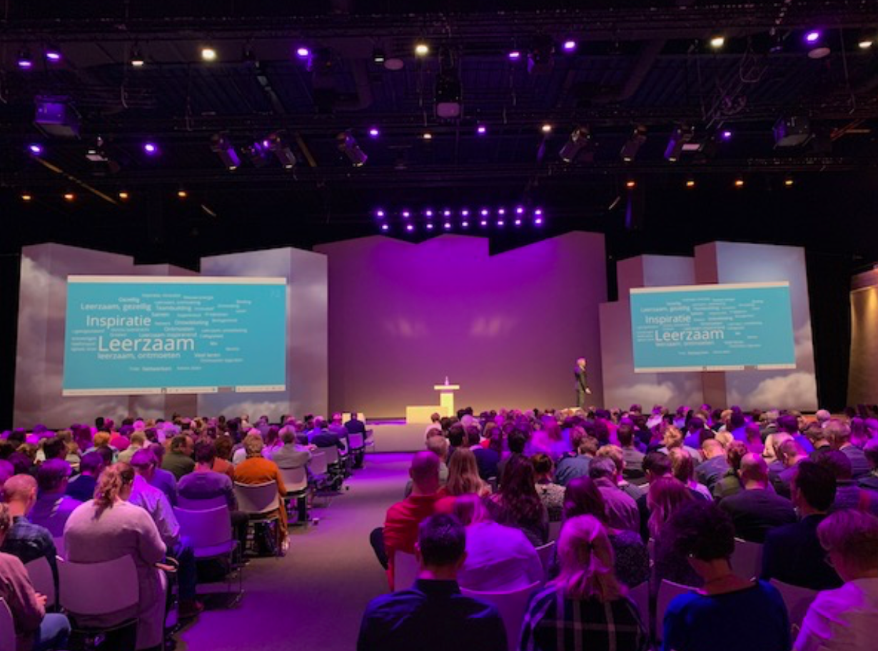 Het publiek zit in een grote zaal, met het gezicht naar een podium met een spreker en schermen met teksten erop, badend in paars licht.