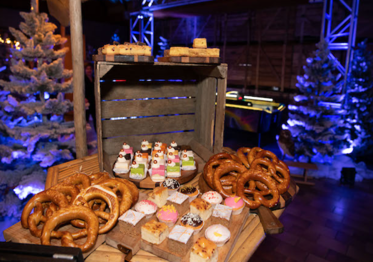 Uitstalling van voedsel met pretzels, gebak en taarten op houten kratten; besneeuwde kerstbomen op de achtergrond.