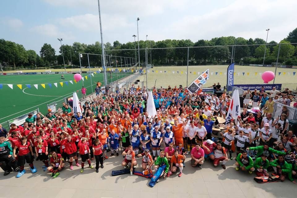 Een grote groep mensen in kleurrijke teamkleding poseert op beton bij een groen veld.