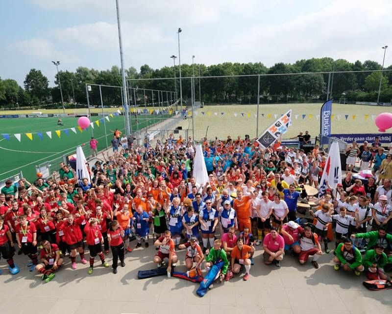 Een grote groep mensen in kleurrijke sportkleding verzamelt zich op een betonnen ondergrond bij een groen veld. Er hangen vlaggen en ballonnen.