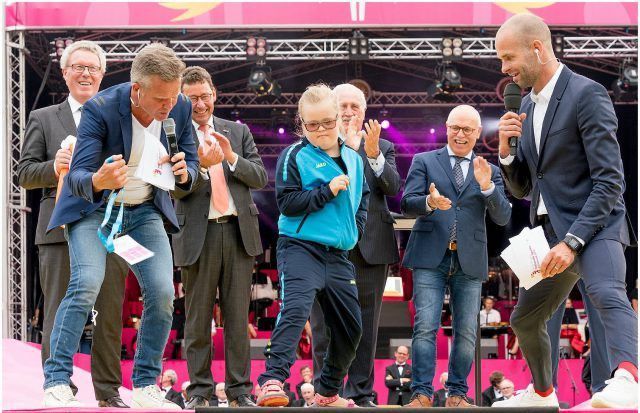 Mensen op het podium: dansende jongens, klappende mannen en microfoons vasthoudend, een podium met een roze achtergrond.
