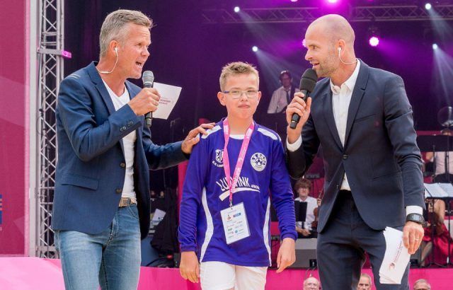 Twee mannen met microfoons flankeren een jongen in een voetbaltenue op een podium; roze achtergrond.