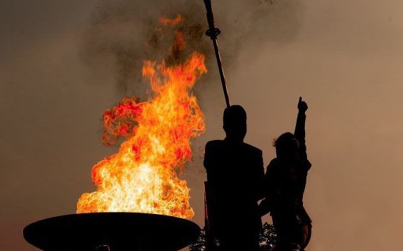 Silhouet van twee personen bij een grote vlam, mogelijk een fakkel aanstekend; zonsondergang.