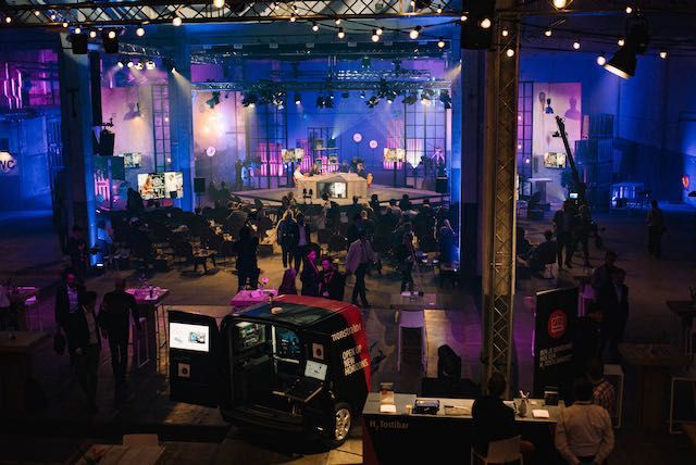 Grote evenementenhal met podium en publiek. Donker, verlicht met blauwe en paarse tinten. Een auto met schermen op de voorgrond.