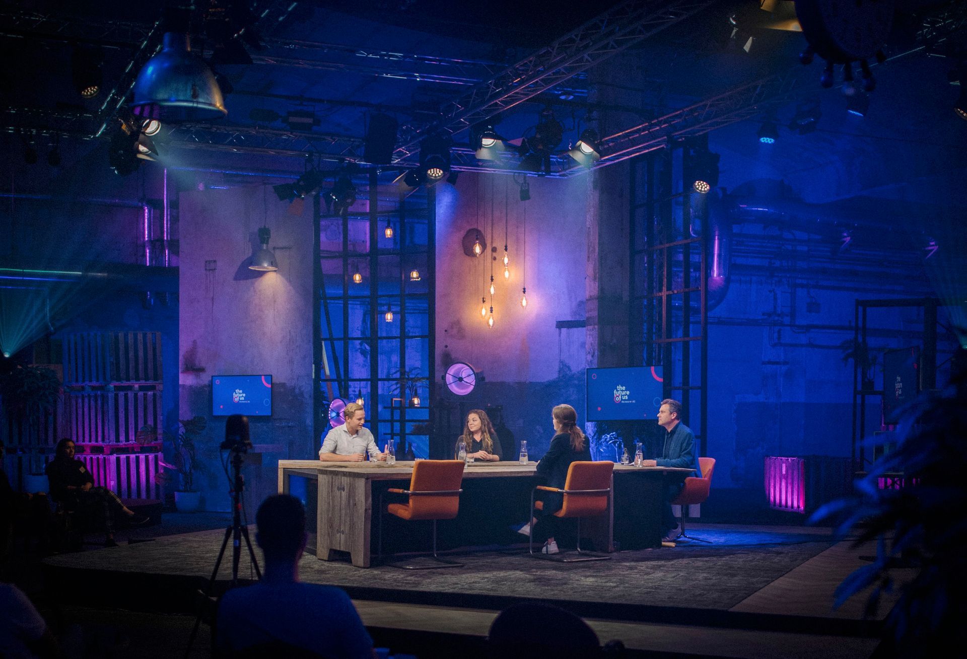 Vier mensen zitten aan een tafel op een podium met een blauw verlichte achtergrond. Een persoon staat tegenover hen.