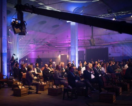 Het publiek zit in een schemerige industriële ruimte en bezoekt een evenement met toneelverlichting en camera's boven hun hoofd.
