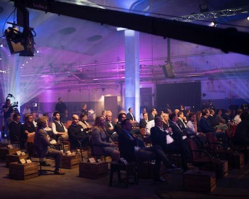 Het publiek zit bij een evenement in een grote, industrieel ingerichte locatie, verlicht met paarse en blauwe podiumlampen.