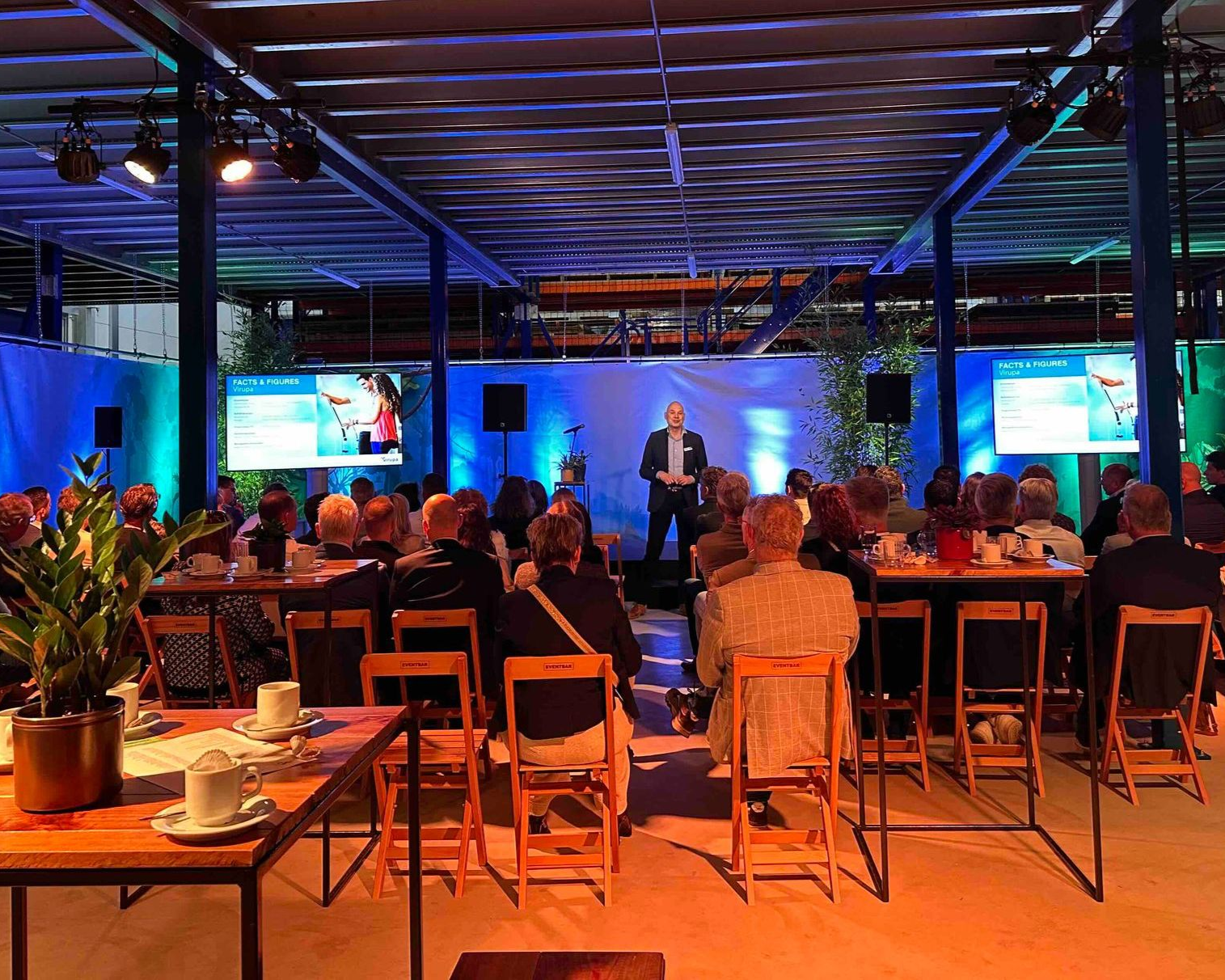 Man geeft op het podium een ​​presentatie aan publiek tijdens een evenement. Schermen, blauwe en groene verlichting, tafels en stoelen.