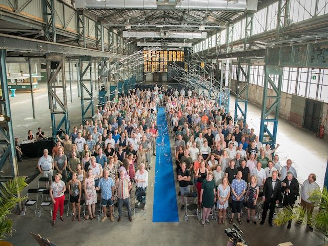 Een grote groep mensen staat in een gebouw in industriële stijl met een gangpad met blauwe loper.
