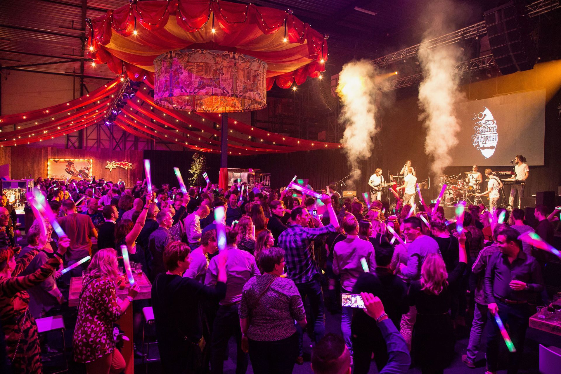 Groot feest in een loods met een band, discolichten en een publiek dat met glowsticks zwaait.